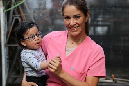 Eine Frau in rosa Kittel hält ein Kleinkind mit Brille auf dem Arm.