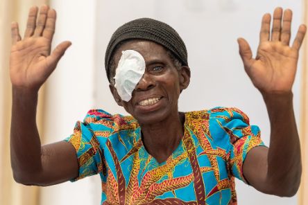 Eine Frau in buntbedrucktem Kleid mit einem Augenverband hält beide Hände hoch und lächelt.