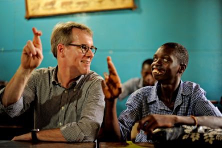 Ein Mann mit Brille und ein afrikanischer Junge sitzen an einem Schulpult und machen das gleiche Fingerzeichen.