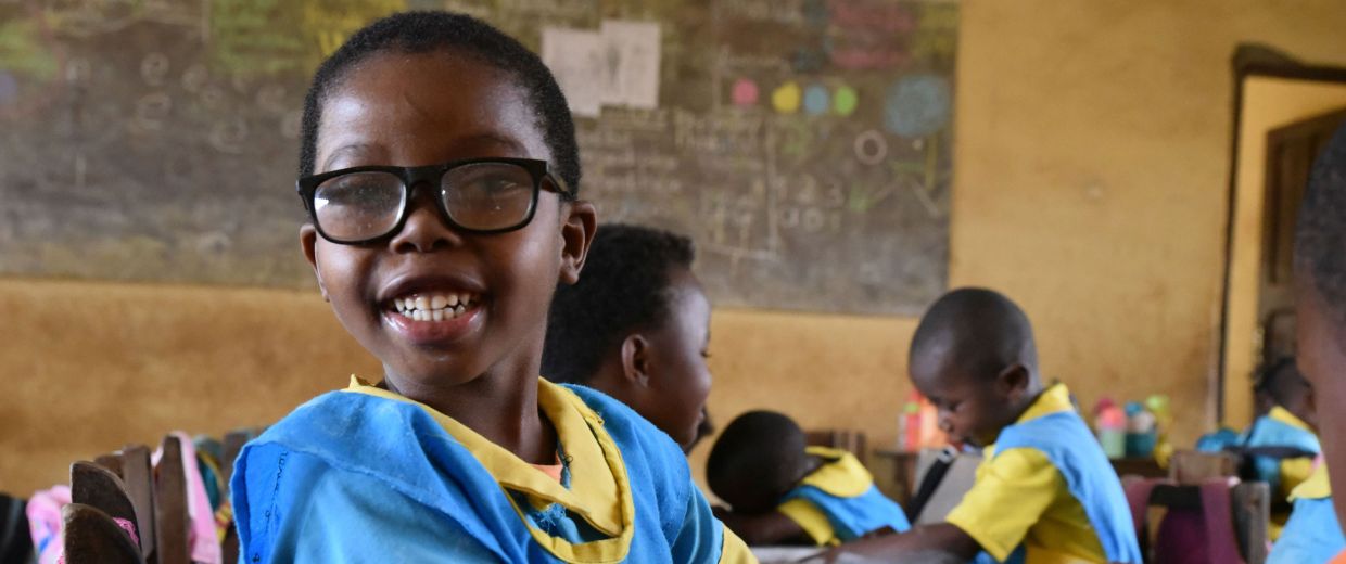 Ein kleines afrikanisches Mädchen mit Brille im Klassenzimmer am Tisch mit anderen Kindern.