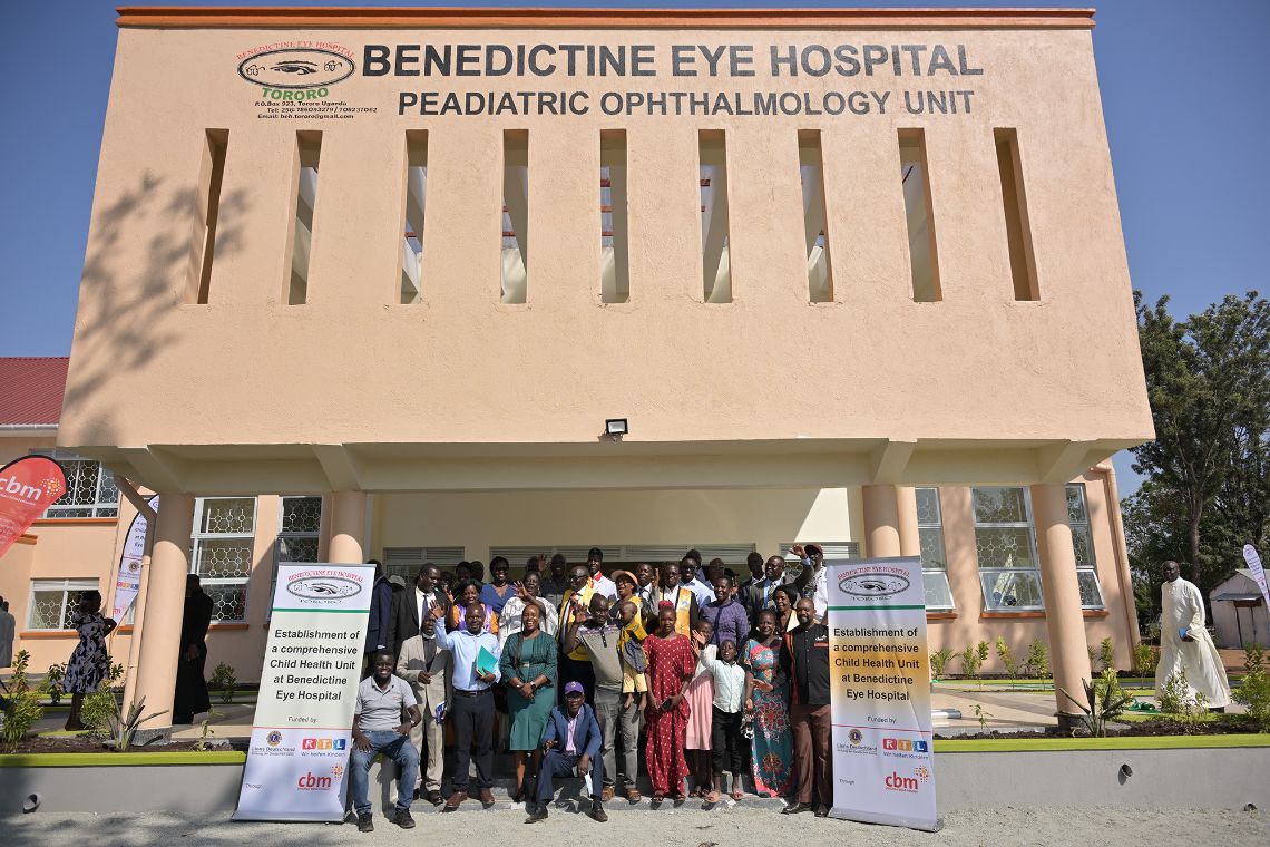 Eine Gruppe von Frauen, Männern und Kindern steht vorm Eingang eines Gebäudes, auf dem steht &quot;Benedictine Eye Hospital – Paediatric Ophthalmology Unit&quot;