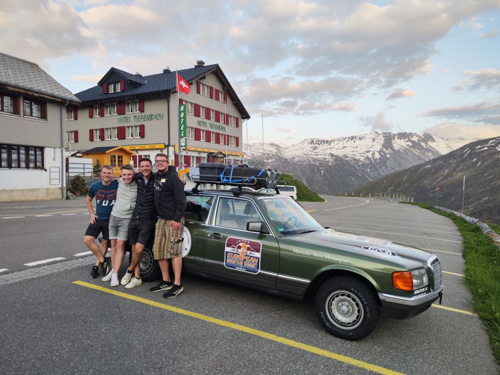 Vier lachende junge Männer stehen an einen alten Mercedes-Benz-Wagen gelehnt, im Hintergrund sind schneebedeckte Berge zu sehen.