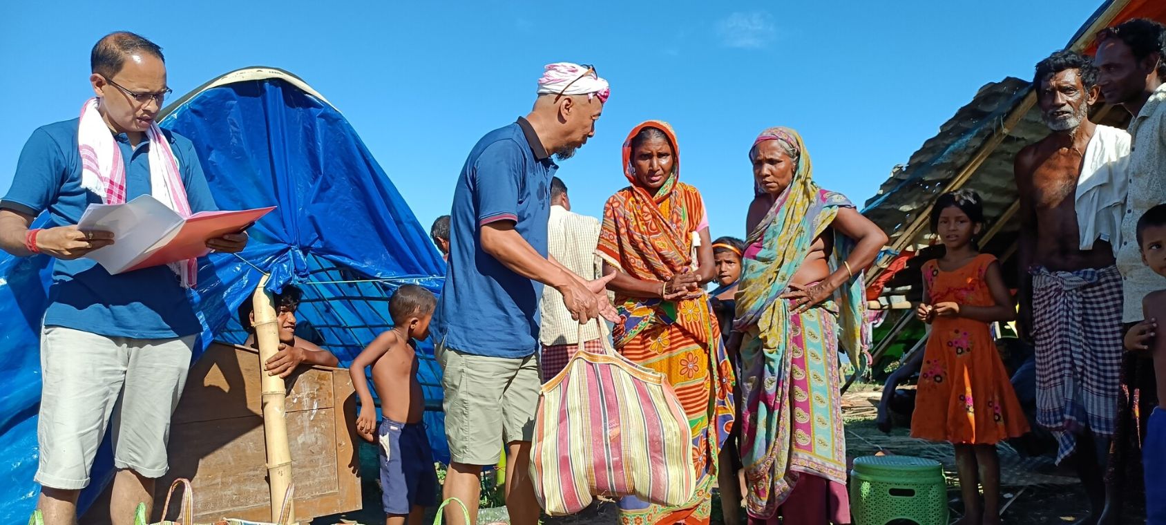 Ein Mann hält eine prall gefüllte Tasche in der Hand. Vor ihm stehen zwei Frauen im Sari, weitere Kinder und Männer stehen in der Nähe.