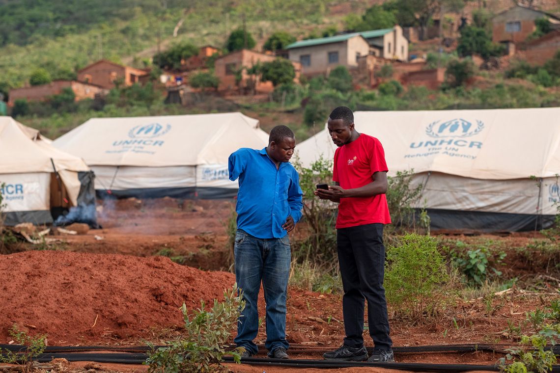 Zwei Männer stehen auf einem Feld vor Zelten mit dem Aufdruck UNHCR. Einem der Männer fehlt ein Arm.