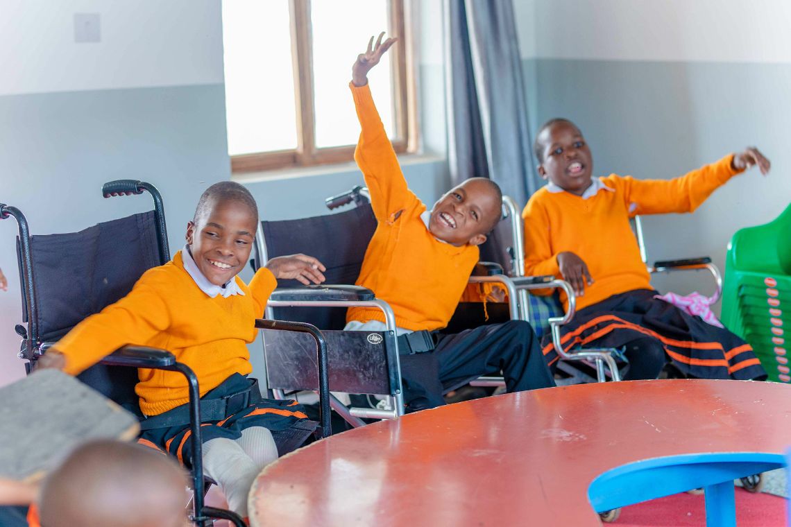 Drei Kinder im Rollstuhl sitzen an einem Tisch im Klassenzimmer. Sie lachen – und zwei von ihnen halten die Hand hoch.