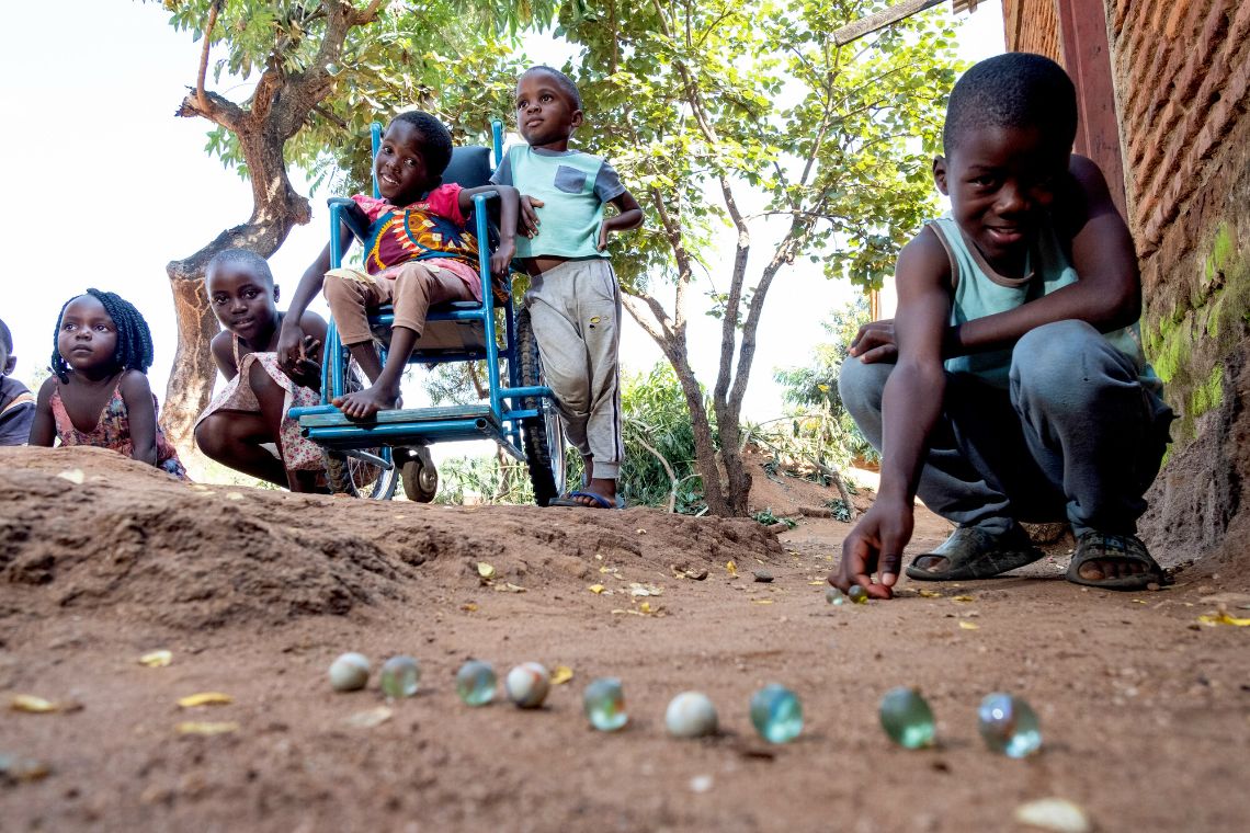 Bunte Glasmurmeln liegen auf dem Erdboden, dahinter sitzt ein Junge in der Hocke. Weitere Kinder, auch eines im Rollstuhl, schauen dem Jungen beim Murmelspiel zu.