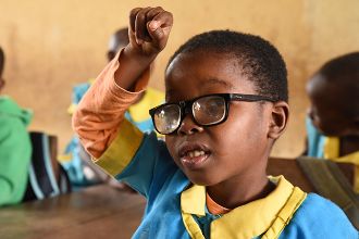 Ein kleines afrikanisches Mädchen mit Brille im Klassenzimmer streckt den Arm hoch.