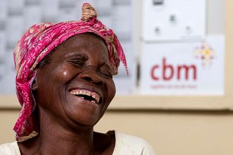 Lachende afrikanische Frau mit buntem Kopftuch, im Hintergrund an einer Pinnwand das CBM-Logo, Spenden zu Weihnachten