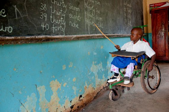 Ein afrikanisches Mädchen sitzt in einem Rollstuhl und deutet mit einem Zeigestock auf eine Schultafel.