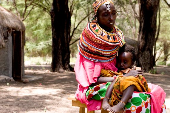 Eine afrikanische Frau sitzt auf einem Hocker und hält ein kleines schwachses Kind im Arm.