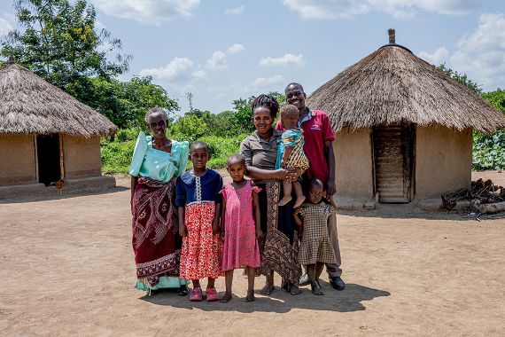 Eine Familie mit Vater, Mutter, Großmutter und vier Kindern steht vor zwei strohgedeckten Rundhütten.