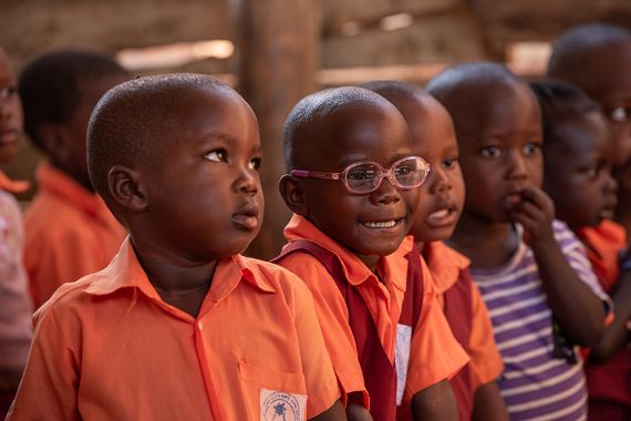 Kinder in Schuluniform sitzen in einer Reihe. Ein Kind trägt eine Brille und lacht.